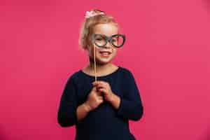 Free photo smiling young blonde girl using paper eyeglasses and looking at the camera over pink wall