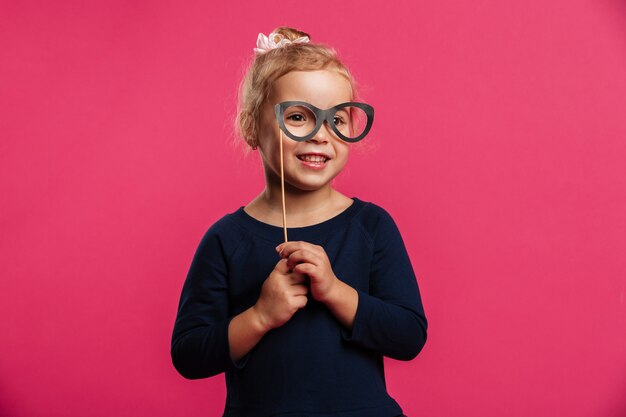 紙の眼鏡を使用して、ピンクの壁の上のカメラを見て笑顔の若いブロンドの女の子