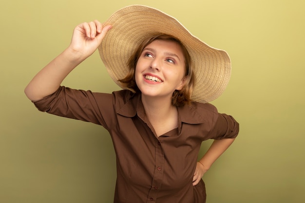 smiling young blonde girl wearing beach hat grabbing hat keeping hand on waist looking at side isolated on olive green wall with copy space