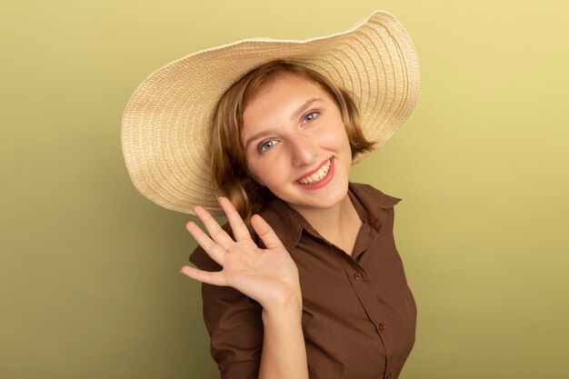 Smiling young blonde girl wearing beach hat standing in profile view looking  waving isolated on olive green wall with copy space