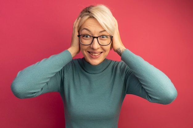 Smiling young blonde girl wearing glasses looking  keeping hands on head isolated on pink wall