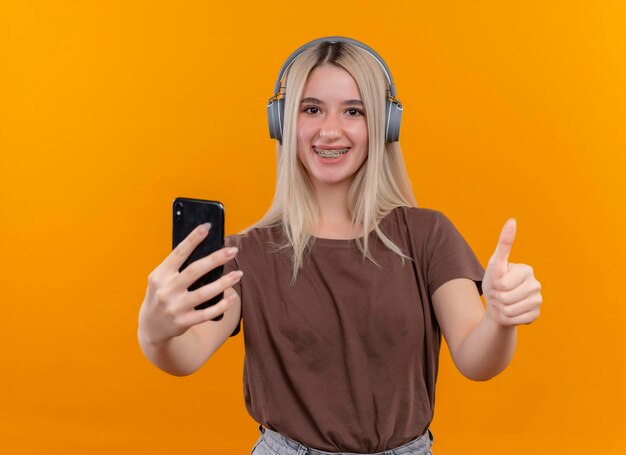 Smiling young blonde girl wearing headphones in dental braces holding mobile phone and showing thumb up on isolated orange space