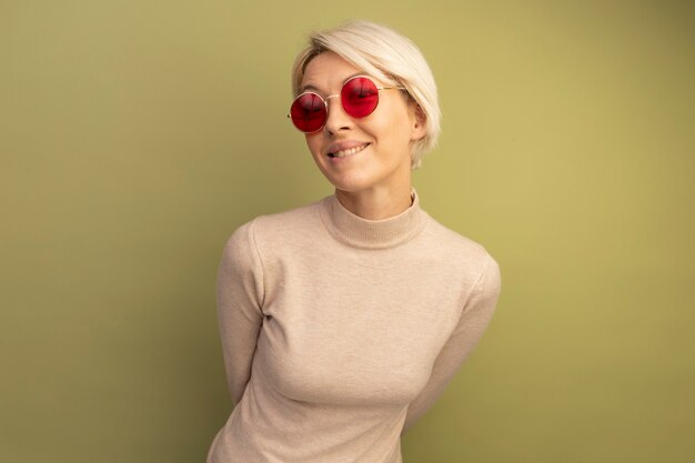 Smiling young blonde girl wearing sunglasses keeping hands behind back looking  biting lip isolated on olive green wall with copy space