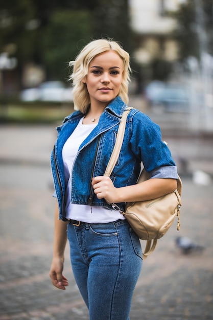 Smiling Young blonde girl woman on streetwalk square fontain dressed up in jeans suite with bag on her shoulder in sunny day