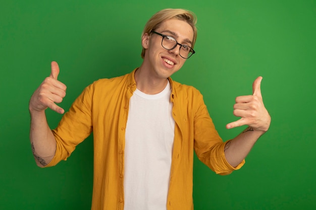 Smiling young blonde guy wearing yellow t-shirt and glasses showing phone call gesture isolated on green