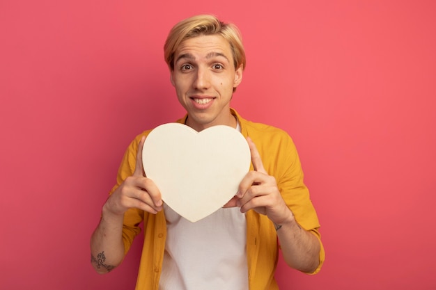 Foto gratuita sorridente giovane ragazzo biondo che indossa la maglietta gialla che tiene la scatola a forma di cuore