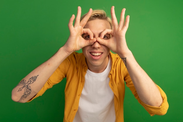 Smiling young blonde guy wearing yellow t-shirt showing look gesture isolated on green