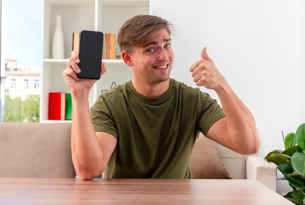 Smiling young blonde handsome man sits at table holds phone and thumbs up inside the living room