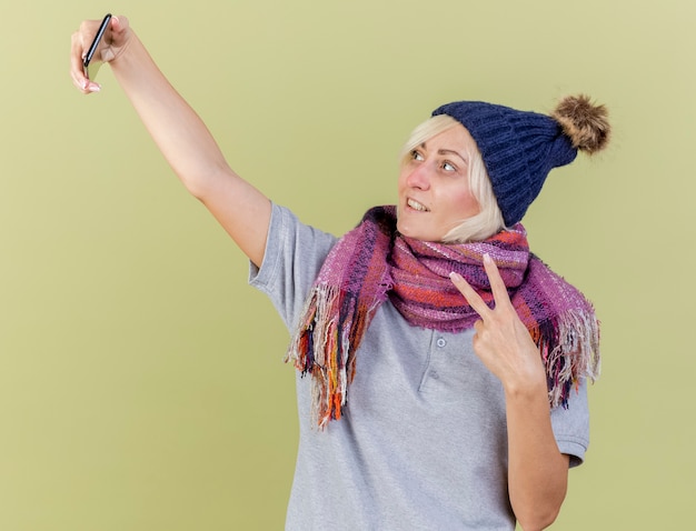 Smiling young blonde ill slavic woman wearing winter hat and scarf gestures victory hand sign looking at phone isolated on olive green wall with copy space