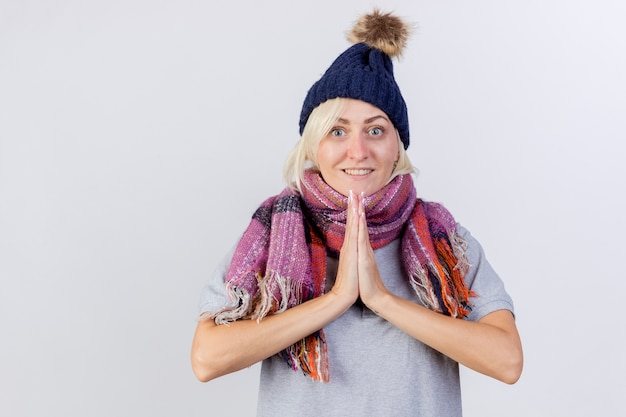 Smiling young blonde ill slavic woman wearing winter hat and scarf holds hands together isolated on white wall with copy space