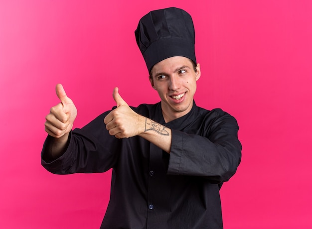 Smiling young blonde male cook in chef uniform and cap looking at side showing thumbs up isolated on pink wall