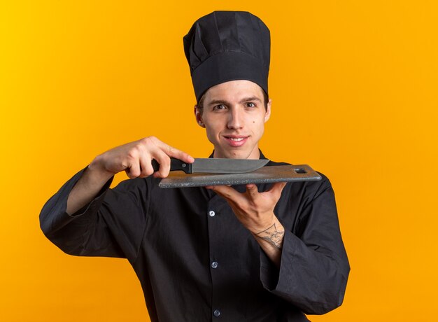 Smiling young blonde male cook in chef uniform and cap touching cutting board with knife looking at camera isolated on orange wall