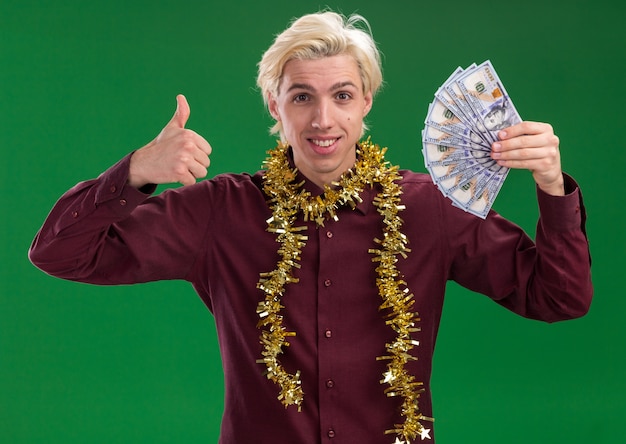 Smiling young blonde man wearing glasses with tinsel garland around neck holding money looking at camera showing thumb up isolated on green background