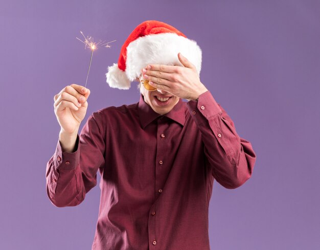 Sorridente giovane uomo biondo che indossa il cappello della santa e occhiali che tengono gli occhi scintillanti vacanza che copre con la mano isolato su sfondo viola