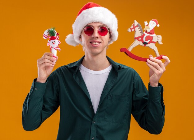 흔들 목마 입상 및 오렌지 배경에 고립 된 카메라를보고 달콤한 지팡이 장식에 산타를 들고 산타 모자와 안경을 쓰고 웃는 젊은 금발의 남자