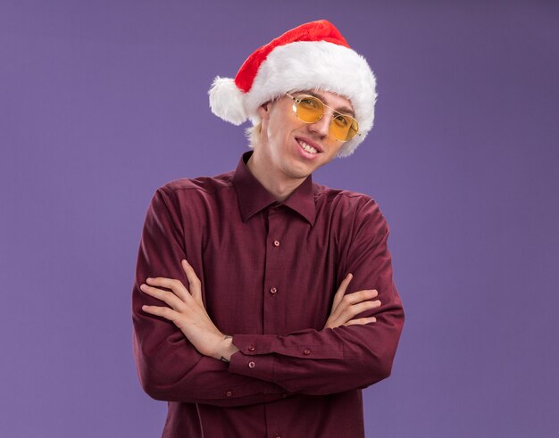 Smiling young blonde man wearing santa hat and glasses standing with closed posture looking at camera isolated on purple background with copy space