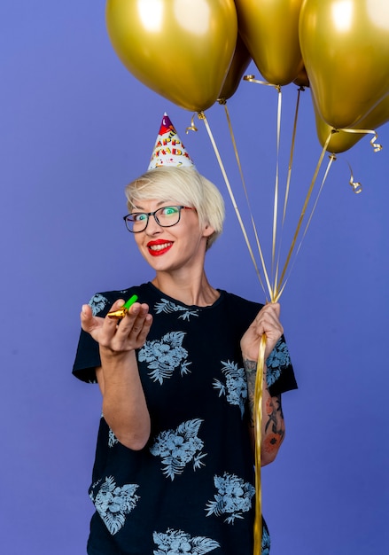 Smiling young blonde party girl wearing glasses and birthday cap holding balloons and party blower looking at camera isolated on purple background
