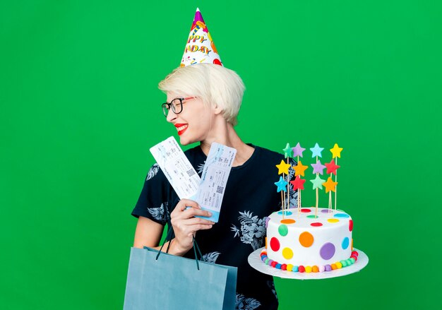 Smiling young blonde party girl wearing glasses and birthday cap holding birthday cake with stars airplane tickets and paper bag with closed eyes isolated on green background with copy space