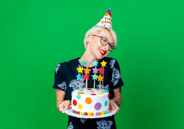 Smiling young blonde party girl wearing glasses and birthday cap holding birthday cake with stars looking at side isolated on green background with copy space