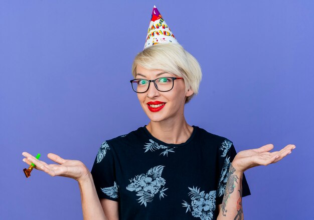 Smiling young blonde party girl wearing glasses and birthday cap holding party blower looking at camera showing empty hands isolated on purple background