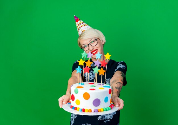 Smiling young blonde party girl wearing glasses and birthday cap stretching out birthday cake with stars looking at camera isolated on green background with copy space