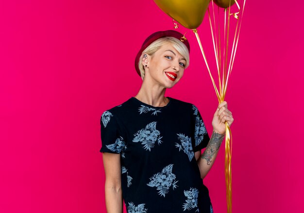 Smiling young blonde party girl wearing party hat holding balloons looking at camera isolated on crimson background with copy space