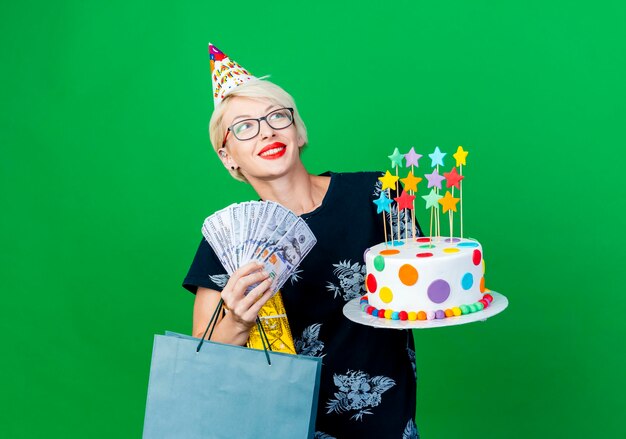 Улыбающаяся молодая светловолосая тусовщица в очках и кепке на день рождения держит торт со звездами, подарочную коробку для денег и бумажный пакет, смотрящую в сторону, изолированную на зеленой стене с копией пространства