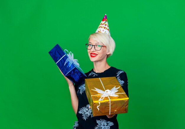 Smiling young blonde party woman wearing glasses and birthday cap holding and stretching out gift box towards front looking at front isolated on green wall with copy space