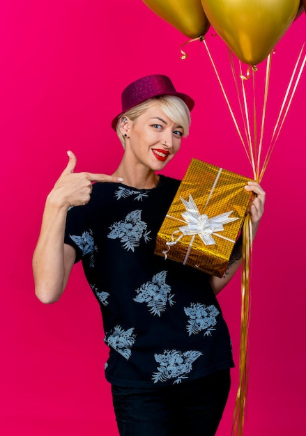 Smiling young blonde party woman wearing party hat holding balloons and gift box pointing at gift box looking at front isolated on pink wall