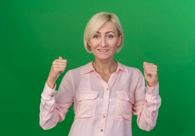 Smiling young blonde slavic woman clenching fists isolated on green background