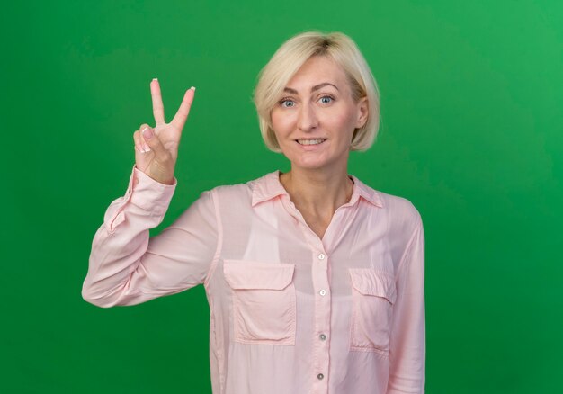 Smiling young blonde slavic woman doing peace sign isolated on green background with copy space