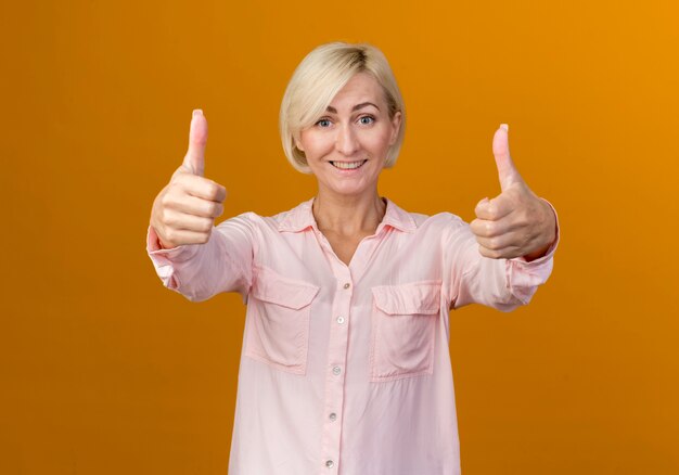 Smiling young blonde slavic woman her thumbs up isolated on orange