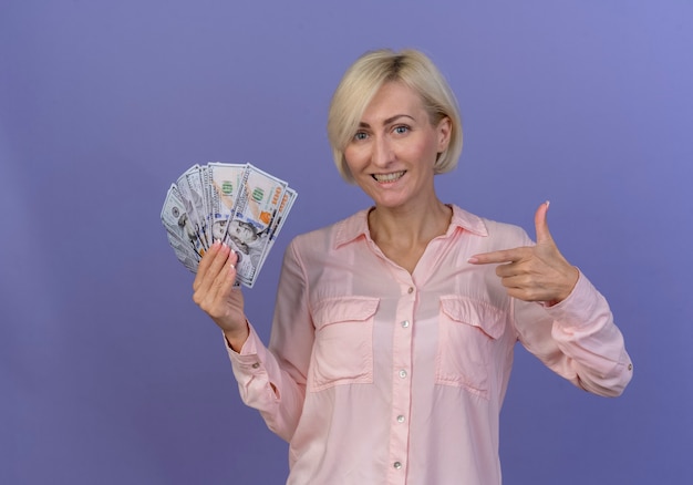 Smiling young blonde slavic woman holding and pointing at money isolated on purple background