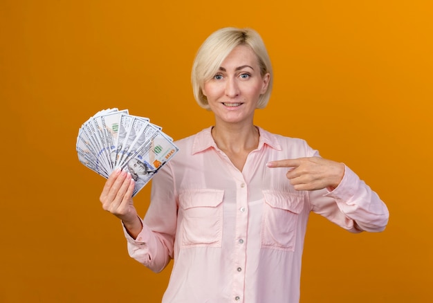 Smiling young blonde slavic woman holding and points at cash isolated on orange wall