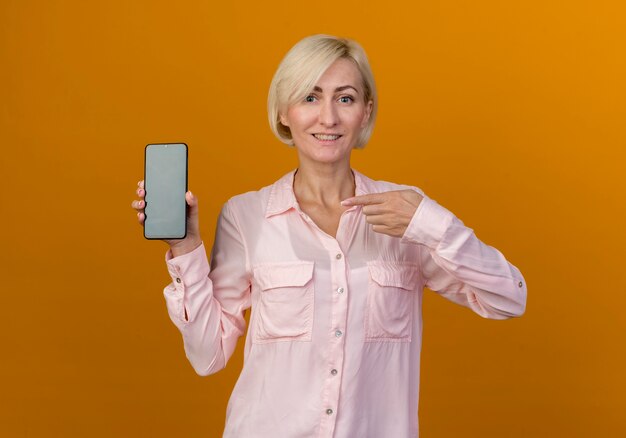 Улыбающаяся молодая белокурая славянская женщина, держащаяся и указывая на телефон, изолирована на оранжевой стене