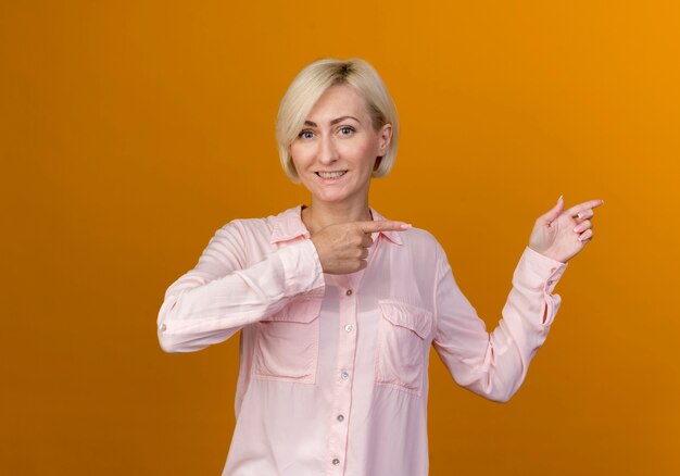 Smiling young blonde slavic woman points at side isolated on orange wall with copy space