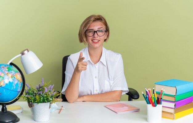 Smiling young blonde student girl wearing glasses sitting at desk with school tools looking and pointing at camera winking isolated on olive green wall