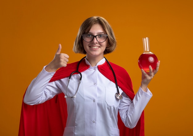 Бесплатное фото Улыбающаяся молодая белокурая женщина-супергерой в красной накидке, в медицинской форме и очках со стетоскопом, держит химическую фляжку с красной жидкостью и показывает палец вверх