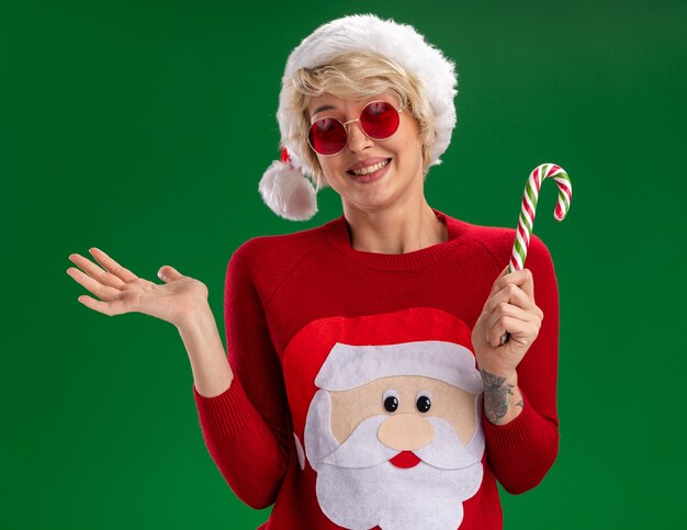smiling young blonde woman wearing christmas hat and santa claus christmas sweater with glasses holding christmas candy cane looking  showing empty hand isolated on green wall