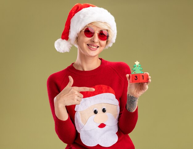 smiling young blonde woman wearing christmas hat and santa claus christmas sweater with glasses holding and pointing at christmas tree toy with date looking  isolated on olive green wall