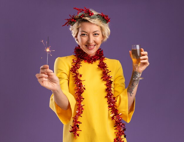 보라색 배경에 고립 된 카메라를보고 휴일 향과 샴페인 잔을 들고 목 주위에 크리스마스 머리 화환과 반짝이 갈 랜드를 입고 웃는 젊은 금발의 여자
