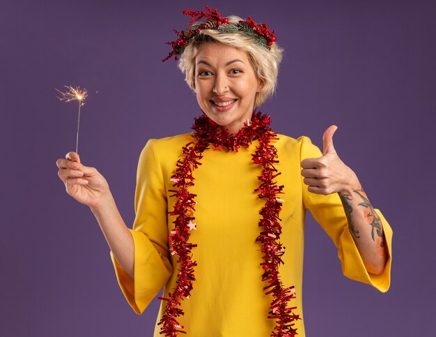 Smiling young blonde woman wearing christmas head wreath and tinsel garland around neck holding holiday sparkler looking at camera showing thumb up isolated on purple background
