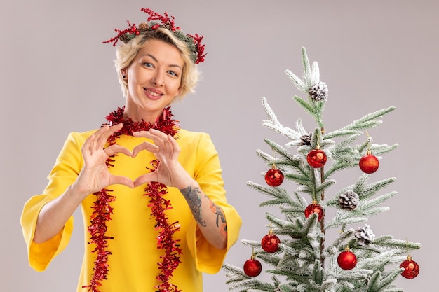 Улыбающаяся молодая блондинка в рождественском венке и гирлянде из мишуры на шее, стоящая возле украшенной елки, глядя в камеру, делает знак сердца на белом фоне