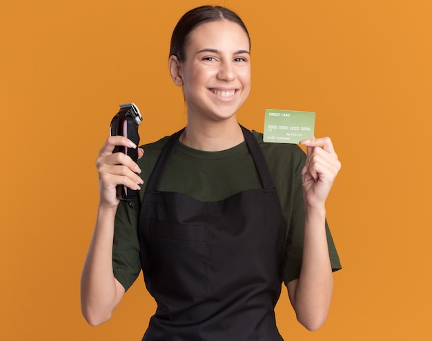 Smiling young brunette barber girl in uniform holding hair clippers and credit card isolated on orange wall with copy space