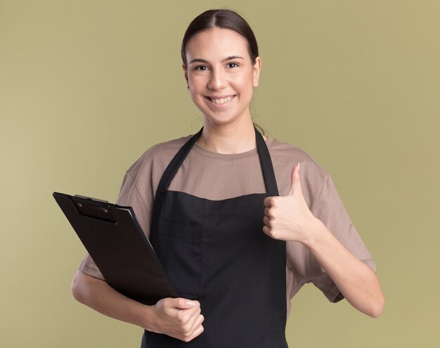 Smiling young brunette barber girl in uniform holds clipboard and thumbs up isolated on olive green wall with copy space