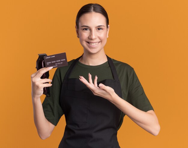 Smiling young brunette barber girl in uniform holds and points at hair clippers and credit card on orange