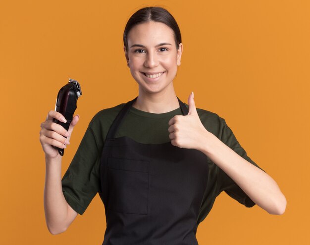 Smiling young brunette barber girl in uniform thumbs up and holds hair clippers isolated on orange wall with copy space