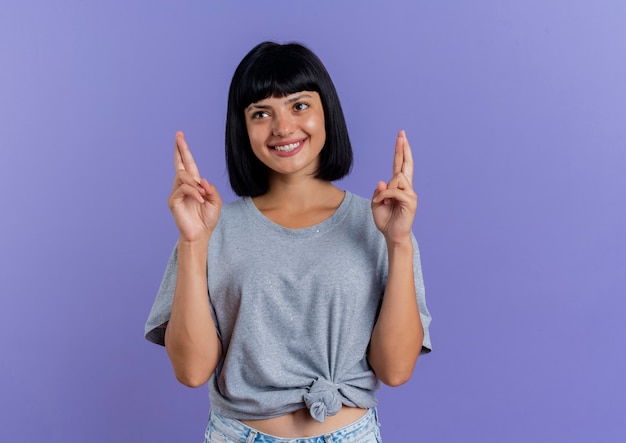 Smiling young brunette caucasian girl crosses fingers looking at side isolated on purple background with copy space