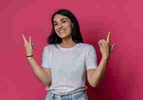 Free photo smiling young brunette caucasian girl gestures horns and gun hand signs isolated on pink wall