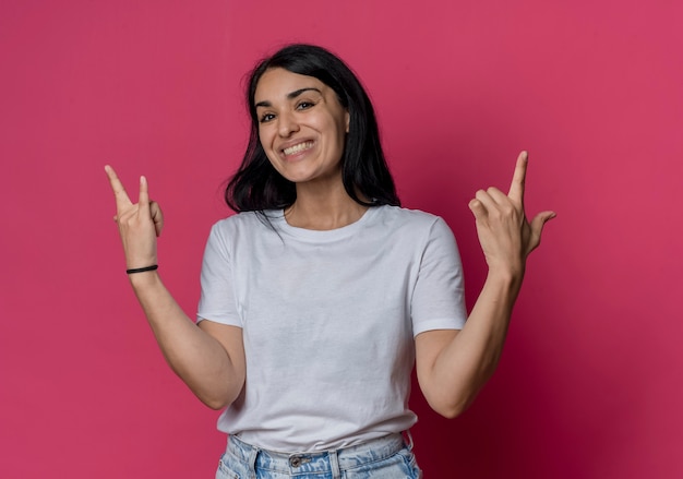 Smiling young brunette caucasian girl gestures horns and gun hand signs isolated on pink wall